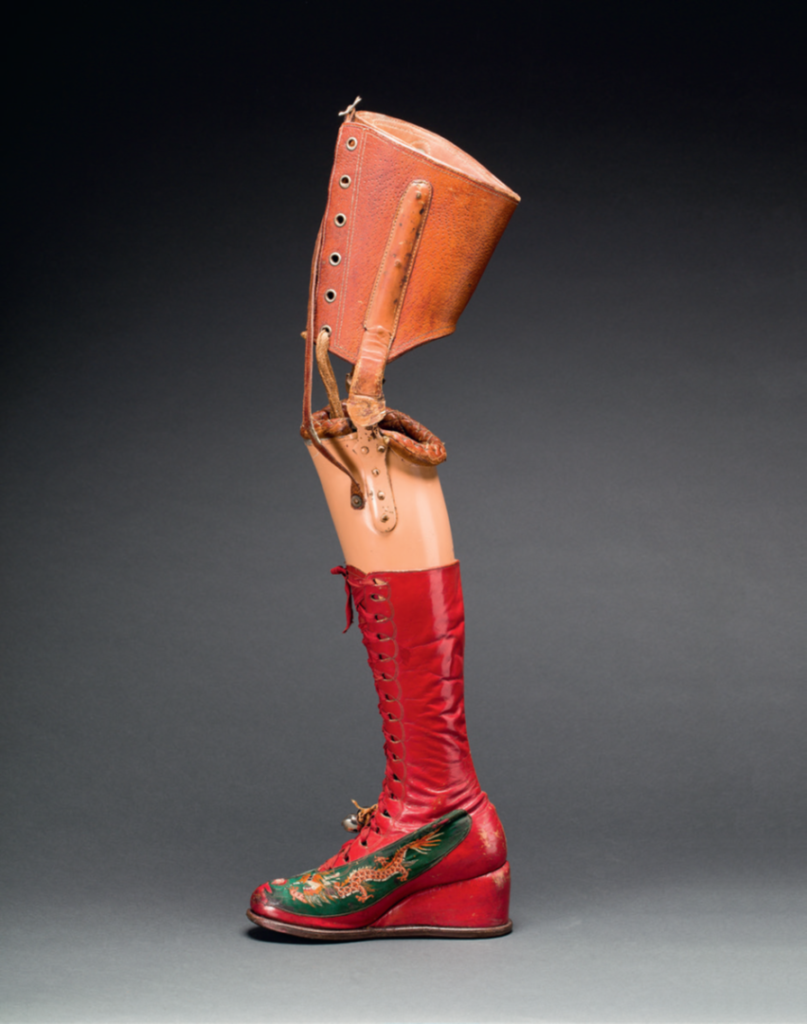 A picture of Frida Kahlo's prosthetic leg, photographed against a grey background. The prosthetic shows a leather part above the knee, where the prosthetic is attached to the amputated leg. The leather part is attached to a plastic, flesh-colored lower leg with two hinges, allowing the leg to bend. The lower leg is wearing a red lace-up boot with a wedge. On the side of the foot Kahlo painted an orange Chinese dragon on top of a green background. Two metallic bells are hanging from a salmon-colored ribbon, attached to the shoelaces at the height of the ankle.